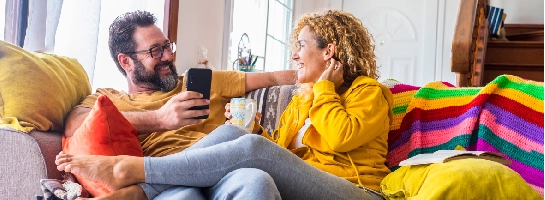 Een stel dat lachend met elkaar op de bank zit met de telefoon in de hand.